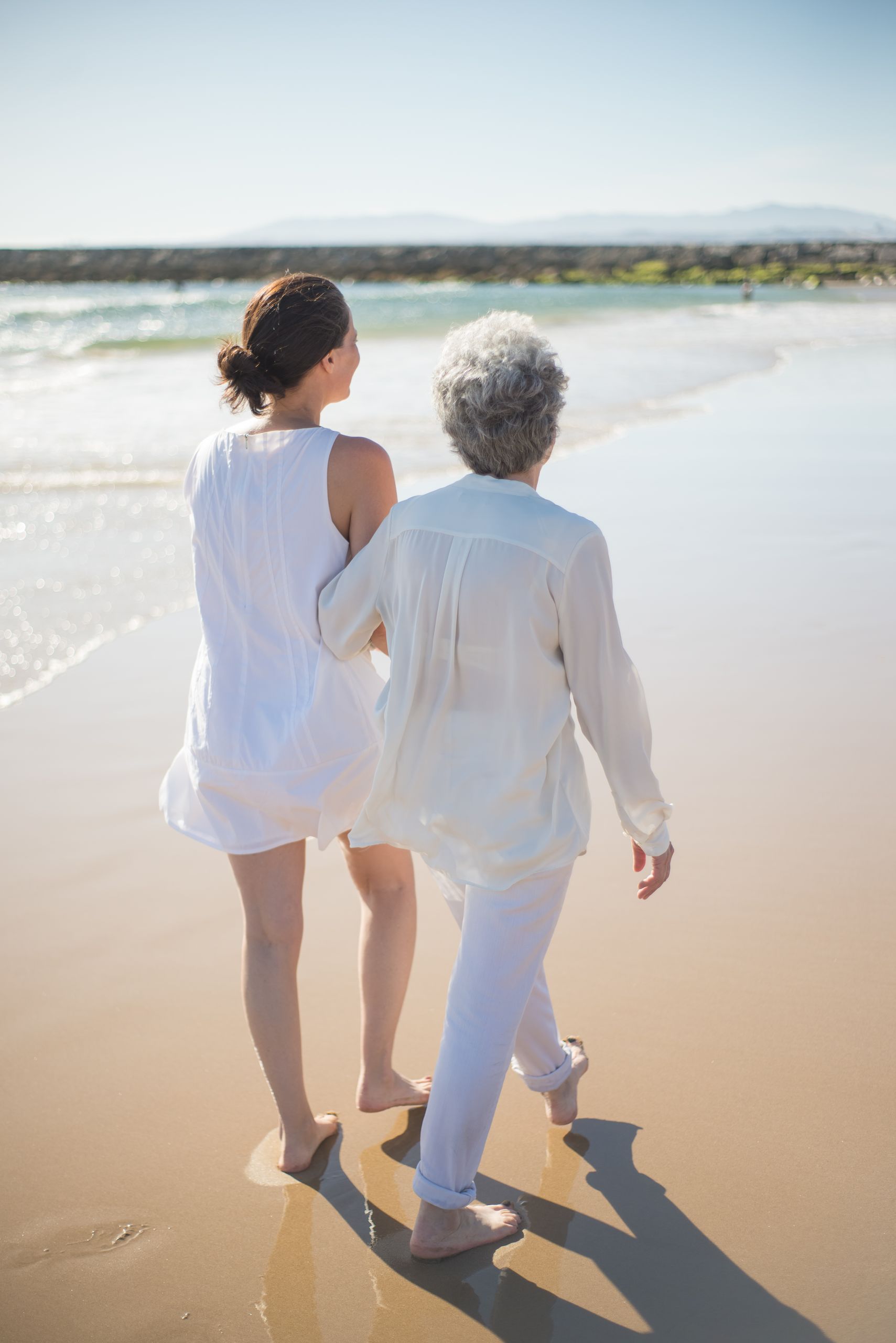 A senior woman and her adult daughter walk barefoot on a sunny beach, enjoying quality time together.